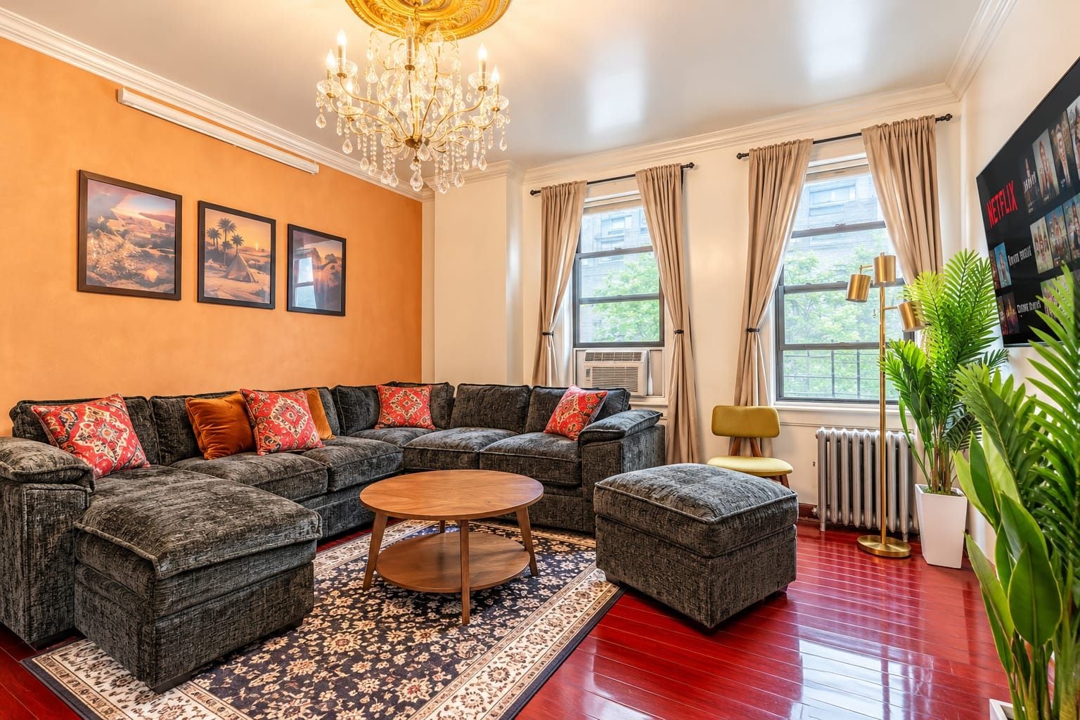 Un salon avec un canapé d’angle, une table basse ronde et un lustre. Il y a deux fenêtres avec des rideaux, un radiateur et une affiche accrochée au mur.