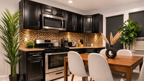 A modern kitchen with black cabinets, stainless steel appliances, a tiled backsplash, and a dining area with a wooden table and chairs. - Lovely 6-bedroom apartment with AC in vibrant Queens (Queens)