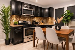 Una cucina moderna con mobili neri, elettrodomestici in acciaio inossidabile, zoccolo a mosaico e zona pranzo con tavolo e sedie in legno.