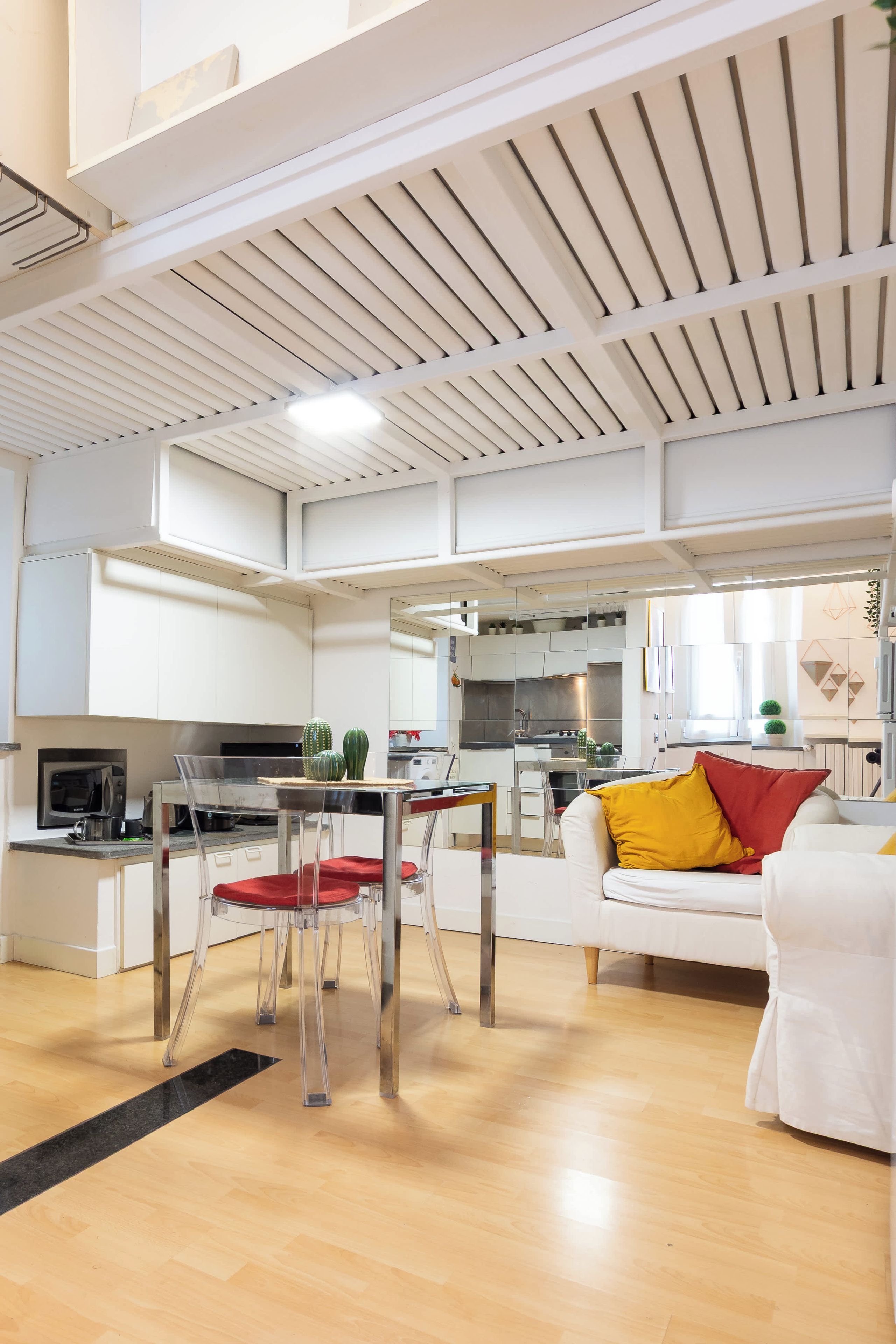 Une cuisine moderne avec des meubles blancs, un coin repas doté d’une table en verre transparent et un espace de vie avec un canapé blanc et des coussins colorés.