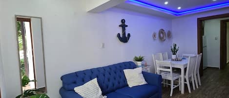 Een woonkamer met een blauwe bank, een eettafel met stoelen, een potplant en een rond vloerkleed.