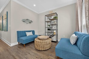 A modern living room with two blue sofas, a wicker ottoman, and a metal shelving unit with decorative items. - The Coastal Drift - Communal Pool (Miramar Beach)