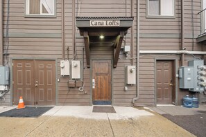 A building with a sign reading 'Cana Lofts' above the entrance. There are two brown doors, a set of electrical boxes, and a water tank on the right side. - Bliss Retreat - Whitefish Studio (Whitefish)