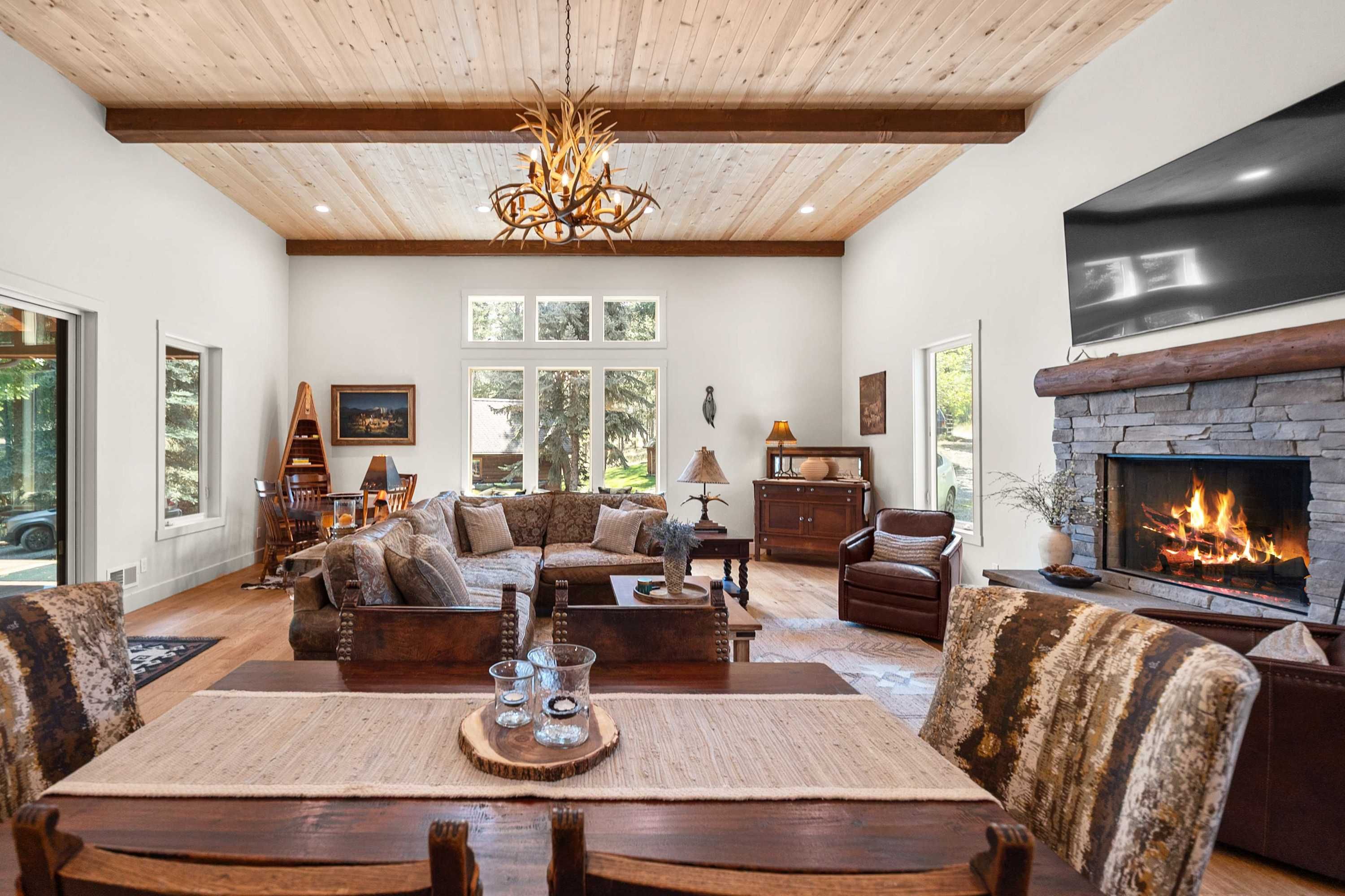 A spacious living room with a fireplace, wooden ceiling beams, and a large window overlooking a forested area.