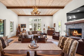 A spacious living room with a fireplace, wooden ceiling beams, and a large window overlooking a forested area. - Whitetail Lodge - Rustic Retreat (Eureka)