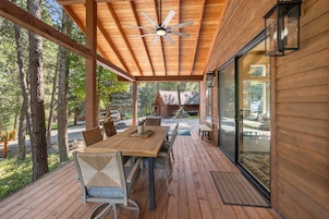 Un área de estar al aire libre techada con mesa y sillas de madera, un ventilador de techo y vistas al bosque circundante.