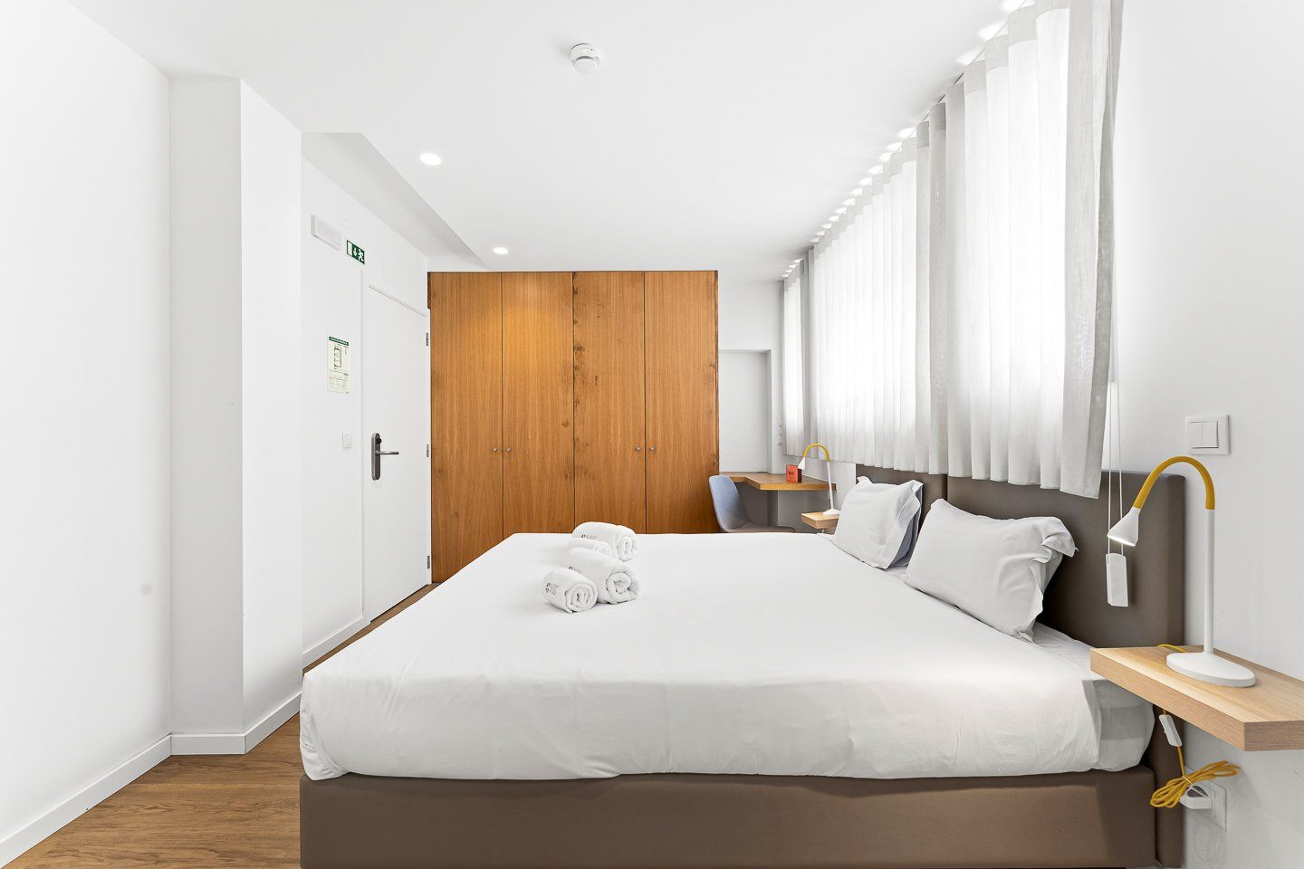 A hotel room with a large bed, a wooden wardrobe, a desk with a chair, and a bedside table with a lamp.