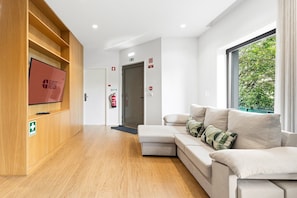 Een hotel lobby met een houten receptiebalie, een flatscreen televisie, een bank met kussens, een brandblusser en een raam met uitzicht op groen.