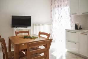 Un comedor con mesa y sillas de madera, un televisor de pantalla plana en la pared, un radiador y una zona de cocina con armarios y una cafetera.