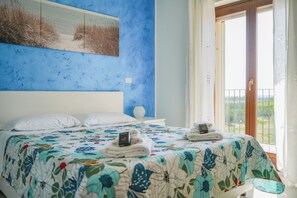 Un dormitorio con cama, almohadas blancas, una mesita de noche con lámpara y vistas al verdor por la ventana.