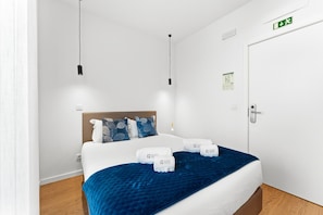 A hotel room with a bed, a nightstand, a wall-mounted light, and a door with an emergency exit sign. - GuestReady - PoetikBlue - Room 7 (Porto)