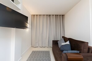 Un salón moderno con un televisor de pantalla plana montado en la pared, un sofá marrón con cojines, una alfombra estampada y un ventanal con cortinas.