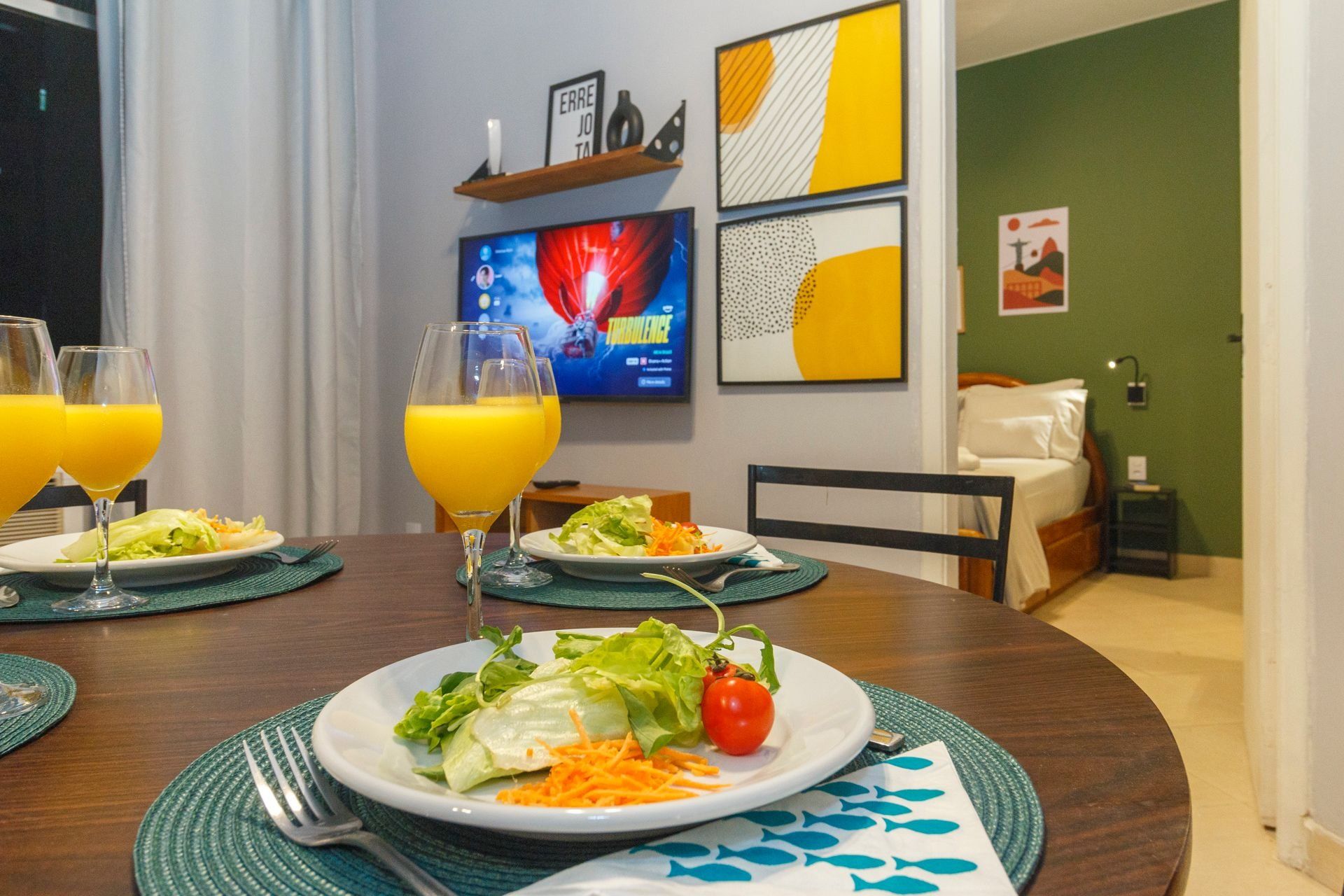 A dining table with plates of food and glasses of orange juice, a television displaying a movie, and a bed in the background.