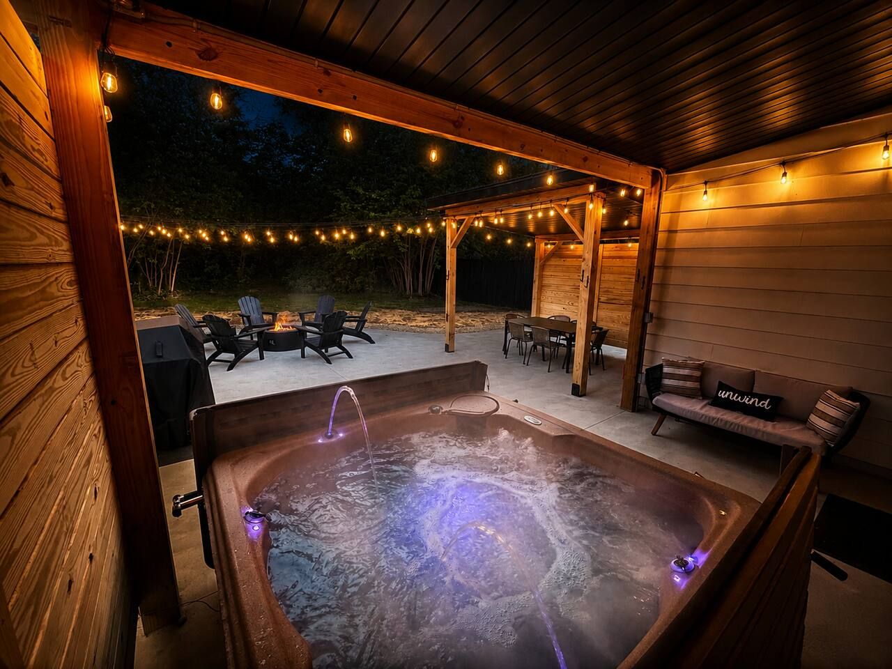 Un bain à remous doté d’une terrasse en bois et illuminé de guirlandes lumineuses, entouré de meubles d’extérieur et d’un espace salon abrité.
