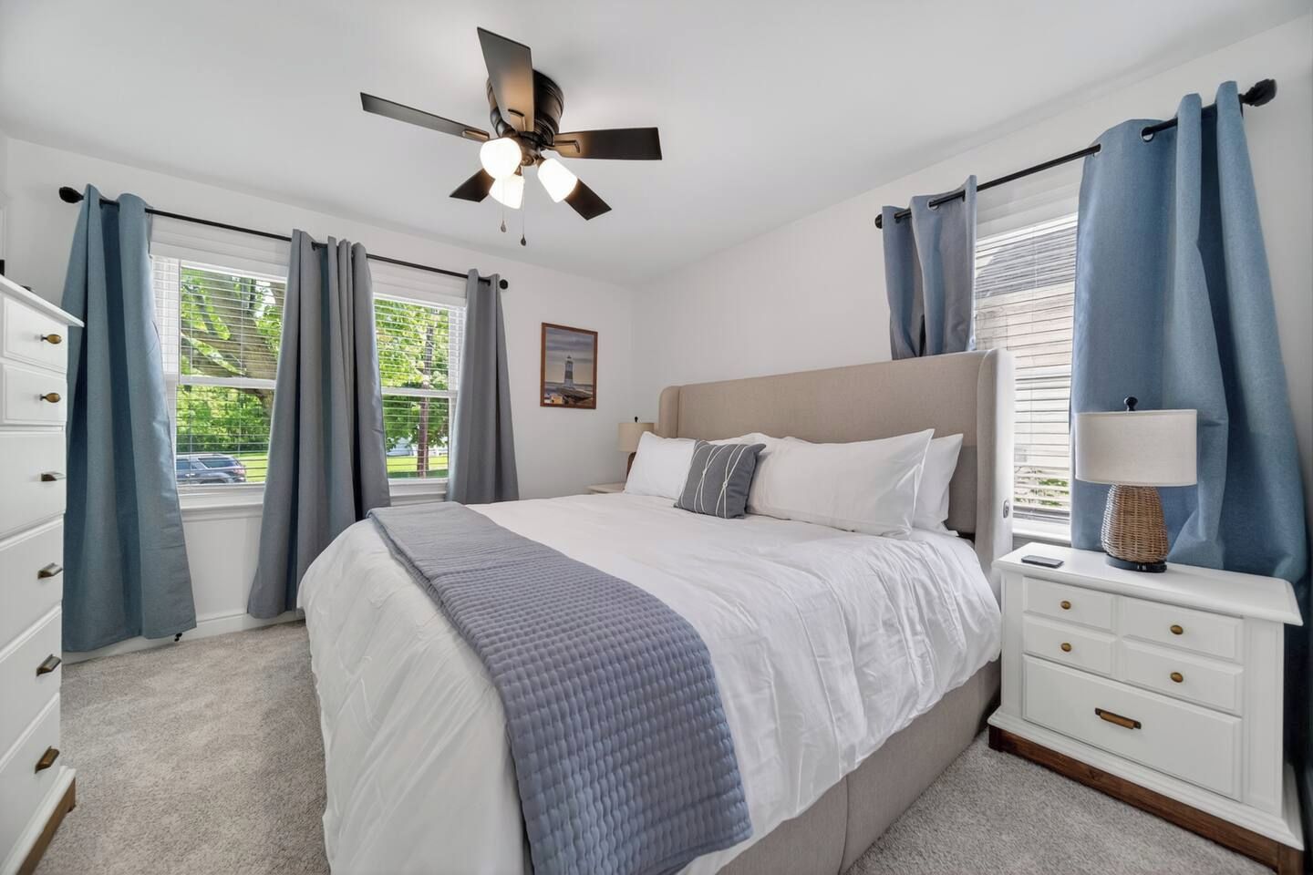 A bedroom with a bed, a nightstand, a dresser, a ceiling fan, and a window with curtains.