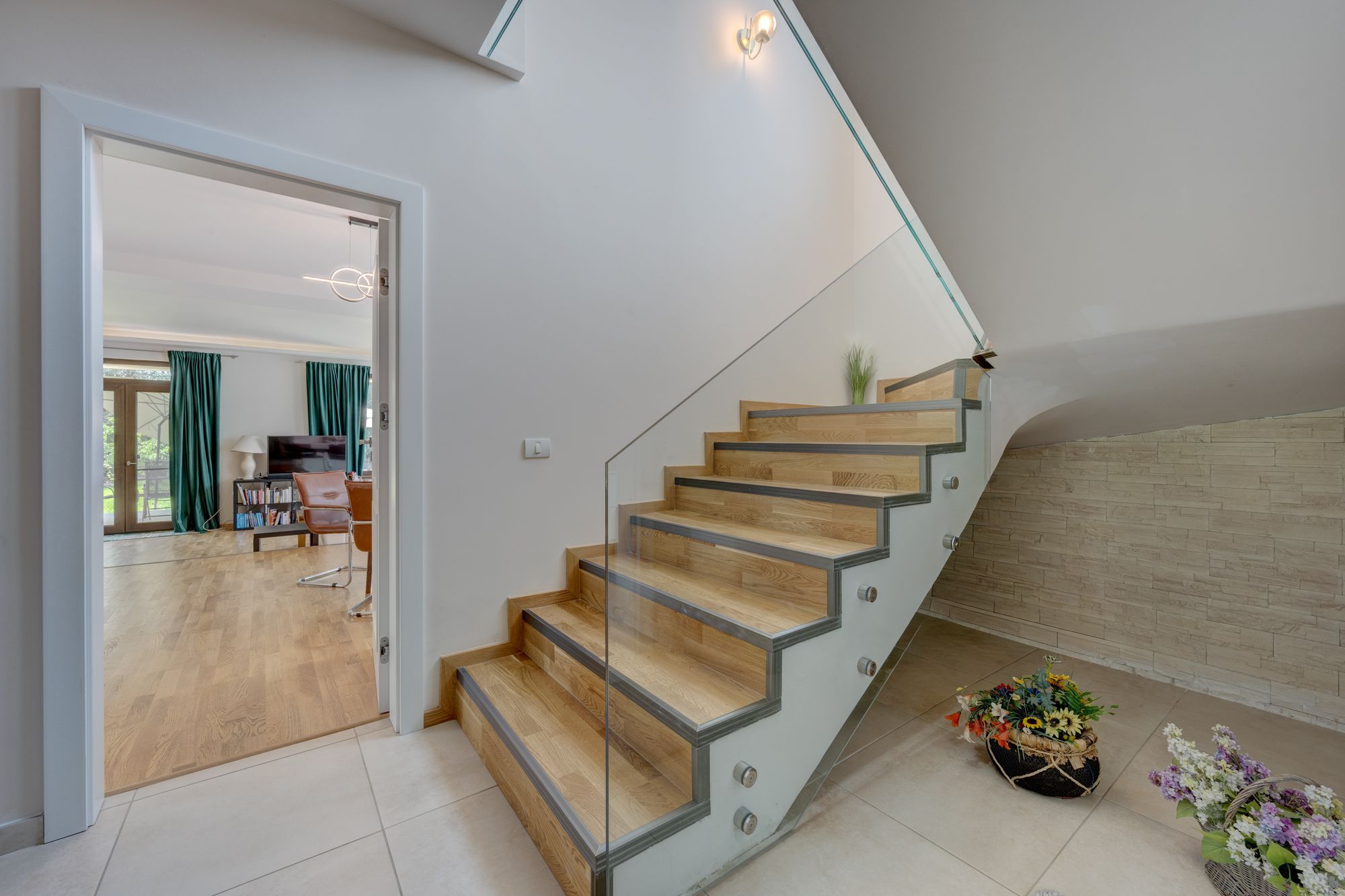 A modern staircase with wooden steps and glass railings, leading to a room with a wooden floor and a view of the outdoors.