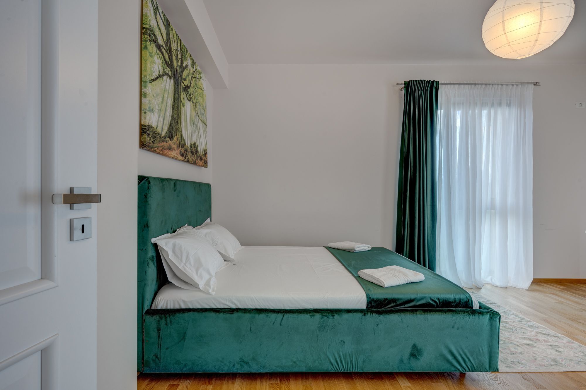 A bedroom with a bed, a painting, a window with curtains, and a door.