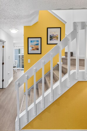 Eine Treppe mit weißen Geländern führt zu einer Empore mit gelben Wänden und zwei gerahmten Bildern.