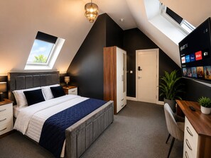 A modern bedroom with a skylight, a bed with a quilted duvet, a desk with a computer, and a TV displaying streaming services. - 4 Bedroom Luxury Apartment in Rushden (Rushden)