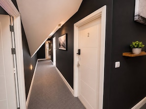 A hallway with a carpeted floor, a door with the number 2, a wall-mounted picture, and a small shelf with a plant.