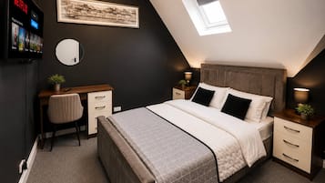 A bedroom with a bed, a desk, a chair, a bedside table, a lamp, a mirror, and a framed picture on the wall.