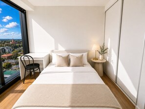 A modern bedroom with a large bed, a desk, and a chair, featuring a city view through a large window. - Suite Dreams - High-Rise City Views (Box Hill)