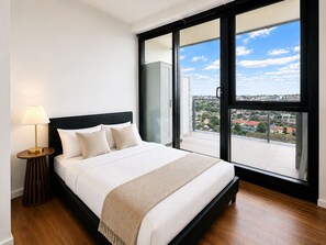 Quarto com cama, abajur e vista para uma área residencial através de grandes janelas.