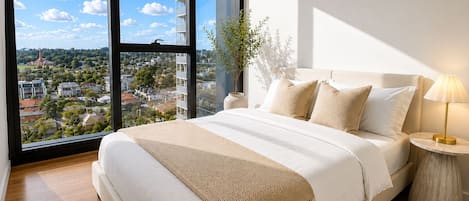 A bedroom with a large window, a bed with white bedding, a bedside table with a lamp, and a plant.