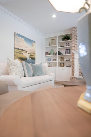 A modern living room with a white sofa, a painting of a landscape, and a brick accent wall. - East Beach Retreat sleeps 12 with a pool (St. Simons Island)
