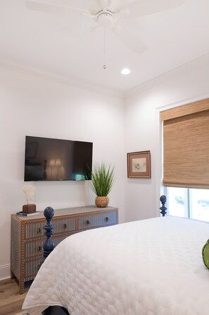 A bedroom with a bed, a television mounted on the wall, a window with a blind, and a decorative plant. - East Beach Retreat sleeps 12 with a pool (St. Simons Island)