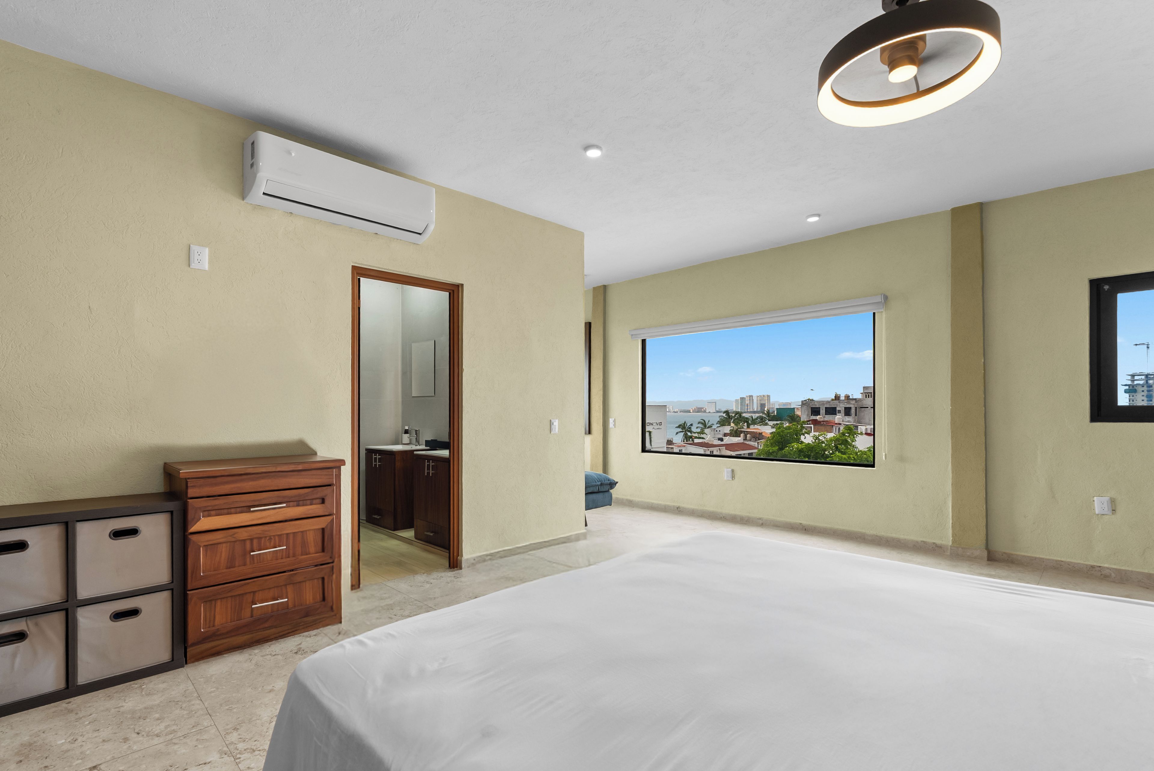 Un dormitorio con una cama grande, un aparador, un equipo de aire acondicionado, una ventana con vista a la ciudad y una lámpara de techo.