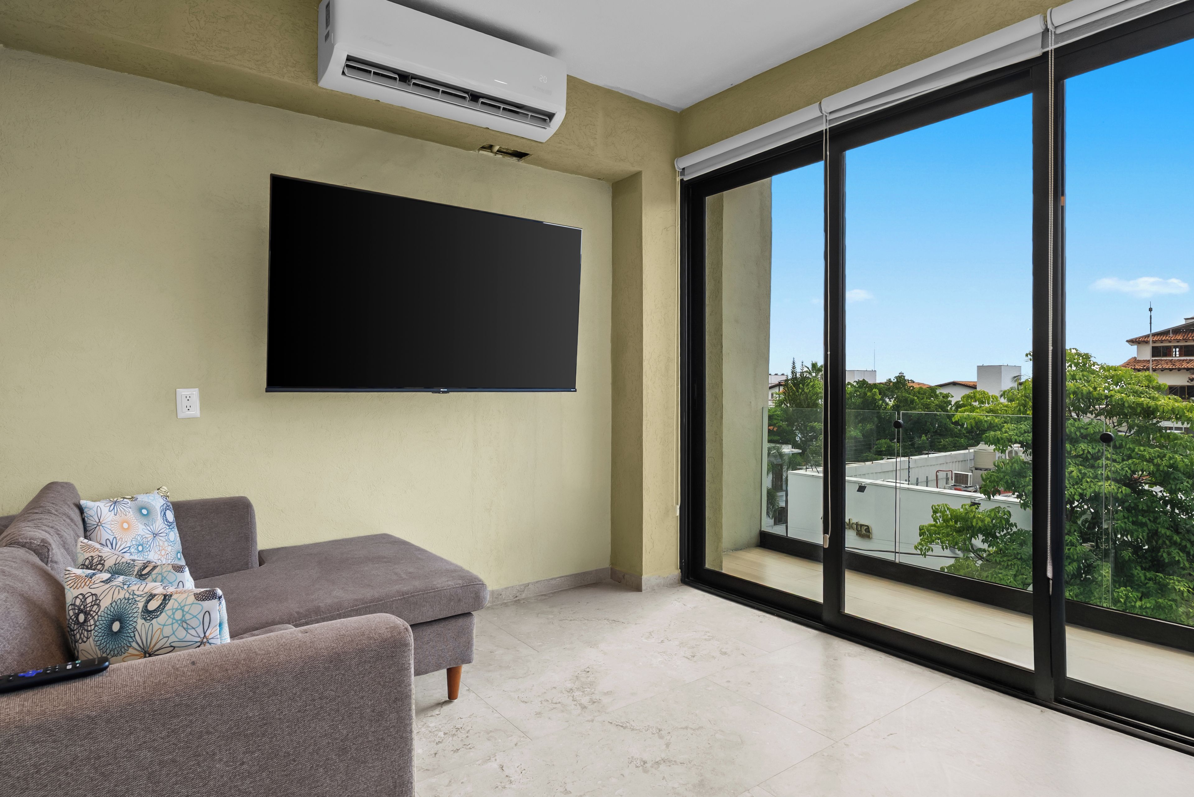 A living room with a flat-screen TV mounted on the wall, a grey sofa with patterned cushions, and a large window offering a view of buildings and trees.