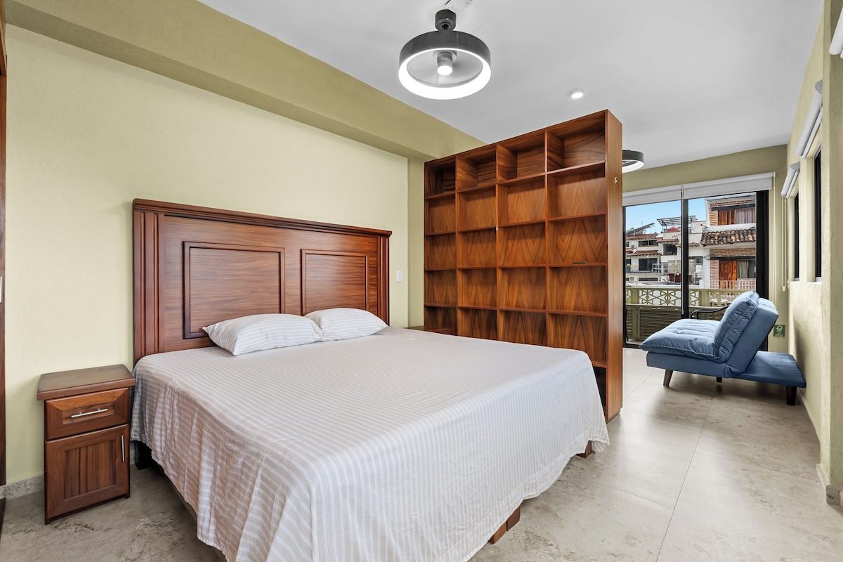 Quarto com uma cama grande, guarda-roupa de madeira, mesa de cabeceira e vista para o exterior.