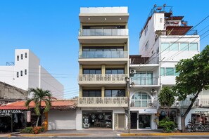 Un edifici de diversos pisos amb un espai comercial a la planta baixa, un balcó amb barana decorativa i un cel blau clar.