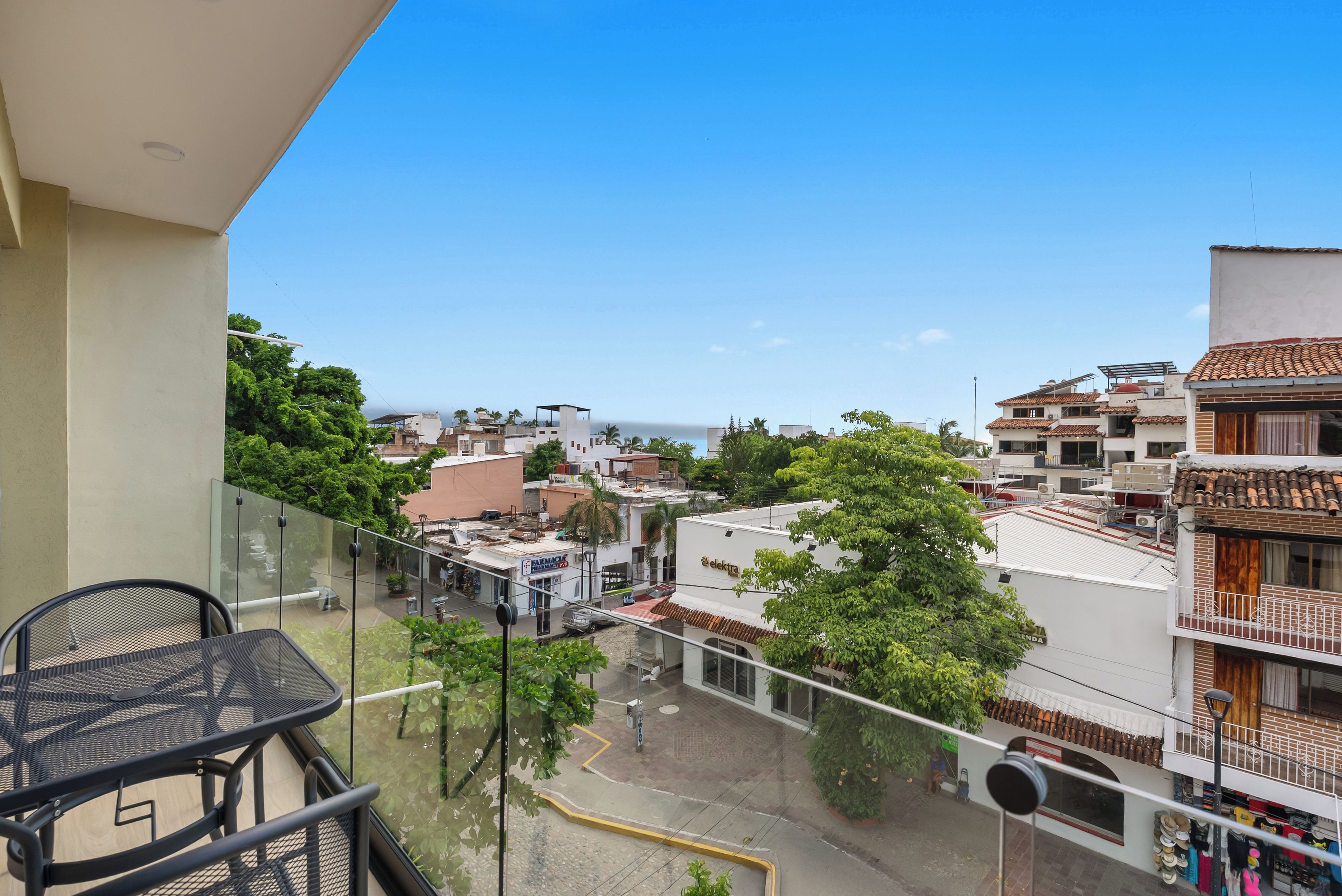 A balcony with a metal table and chairs, overlooking a street with buildings and trees.