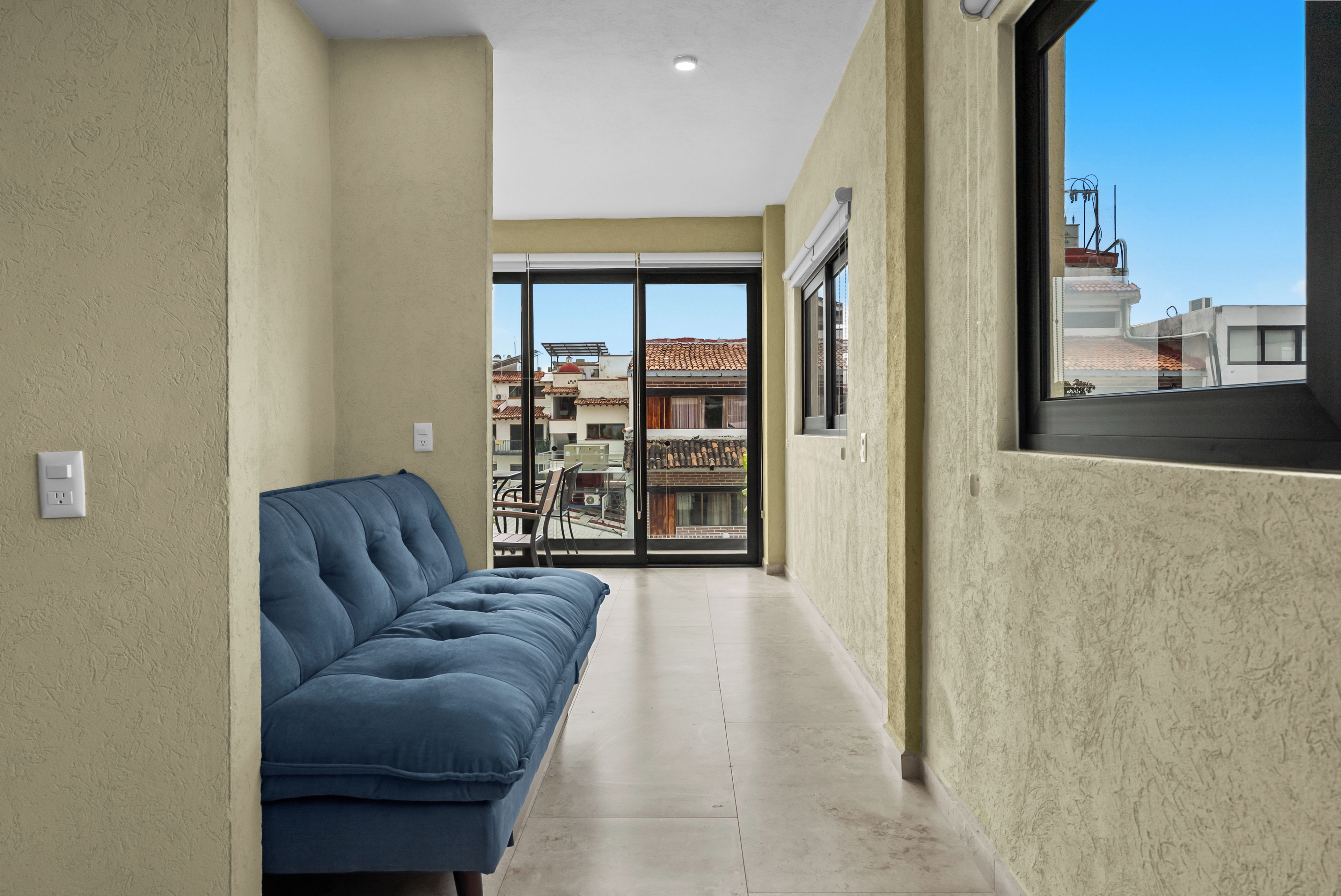 Un pasillo con un sofá azul, un vidrio que da a un área exterior con edificios, y una ventana a la derecha.