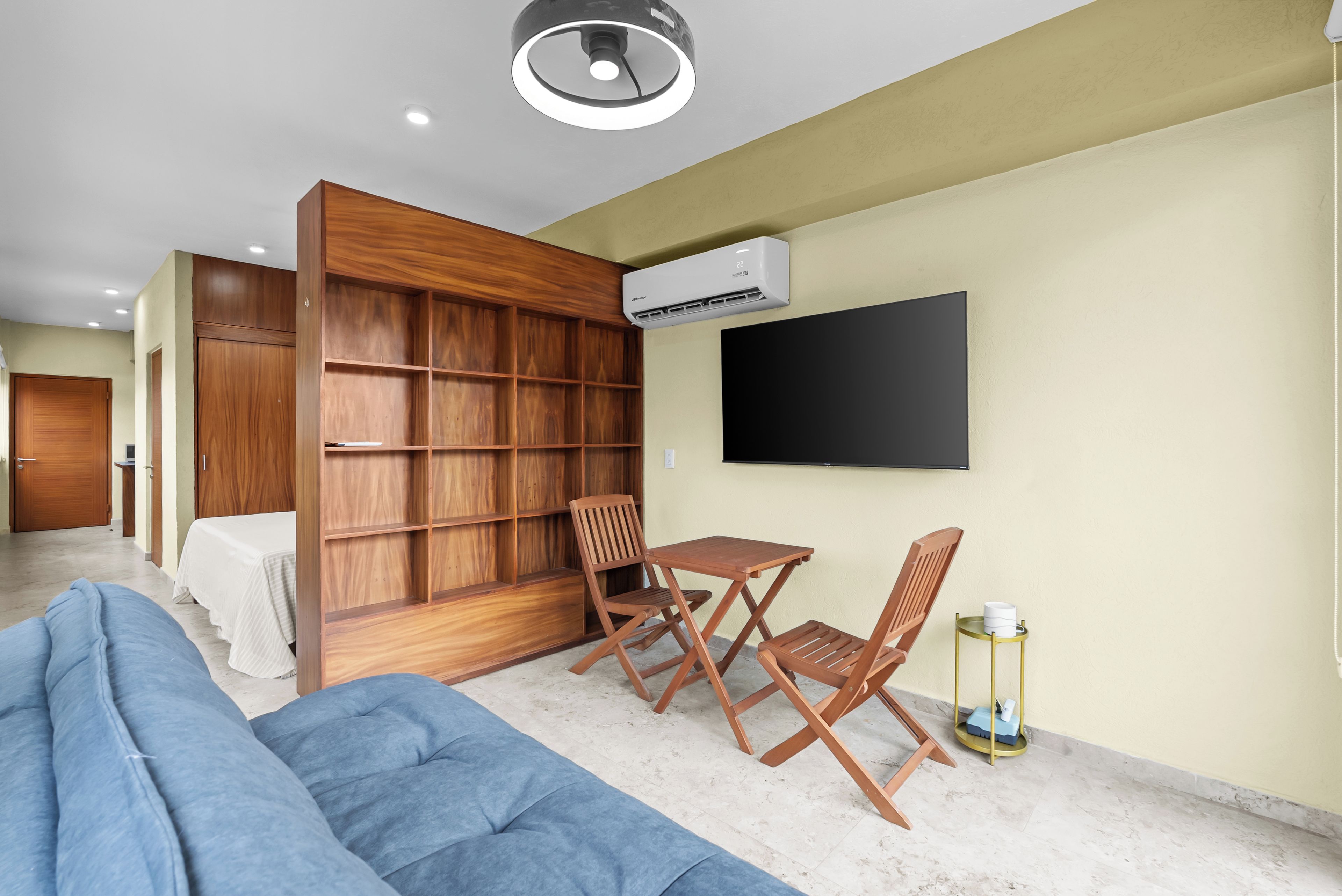 A modern living room with a sofa, a wooden bookshelf, a TV mounted on the wall, and a small table with chairs.