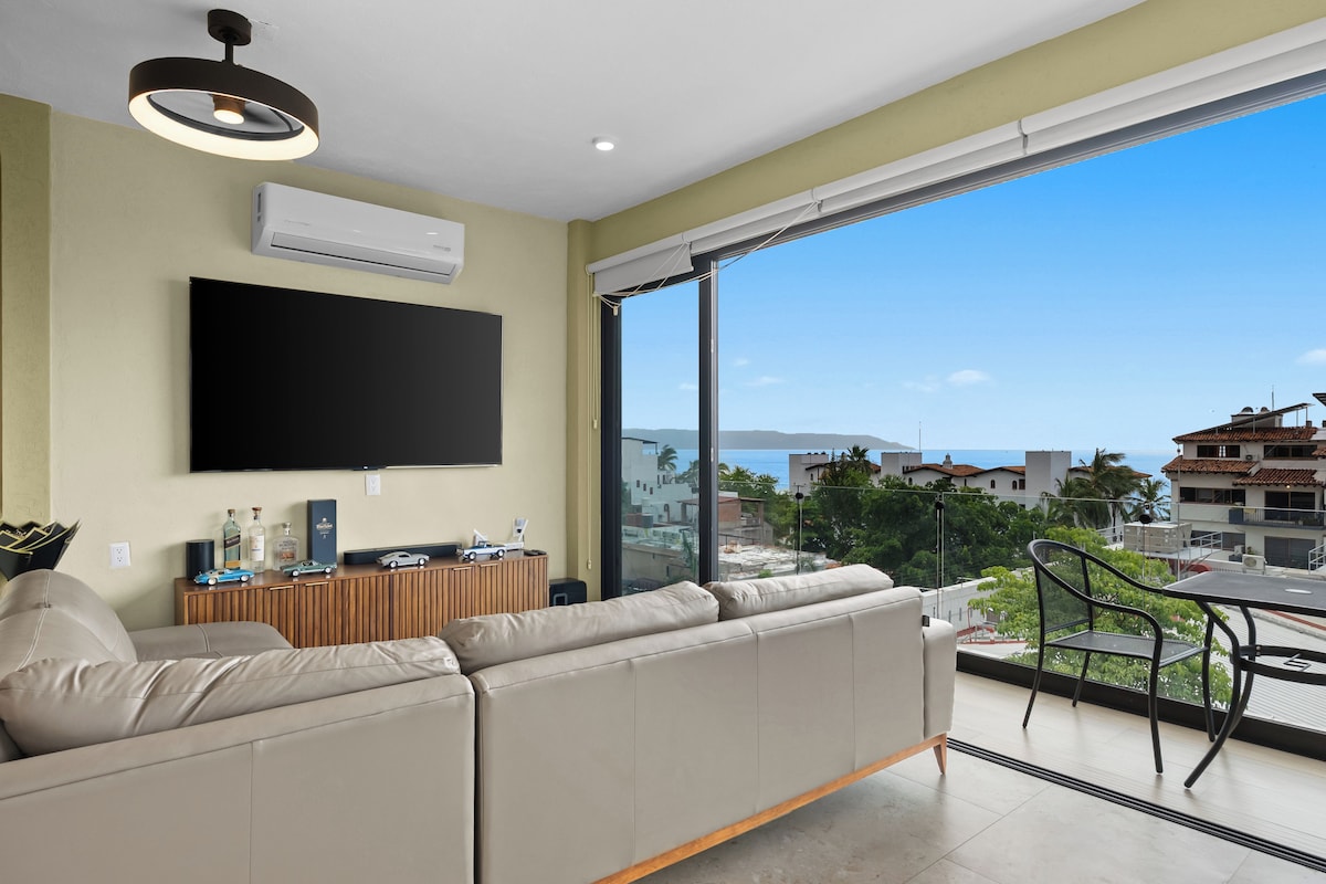 Sala de estar moderna com uma televisão de tela grande, um sofá e vista para a cidade e o mar através de uma janela ampla.