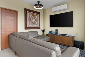 Un saló amb un sofà seccional, una televisió de pantalla plana, una obra decorativa a la paret i un armari de fusta amb objectes petits a la part superior.
