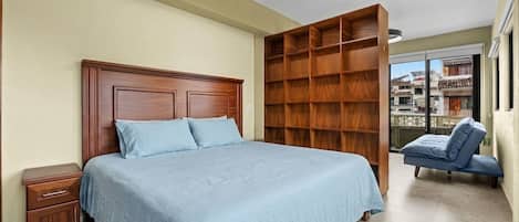 Een slaapkamer met een houten bed, nachtkastje, een inbouwkast met houten planken en uitzicht naar buiten door een groot raam.
