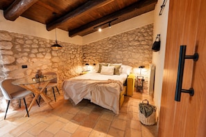 A cozy bedroom with a stone wall, a bed with a white bedspread, a round glass table with chairs, and a wooden ceiling.