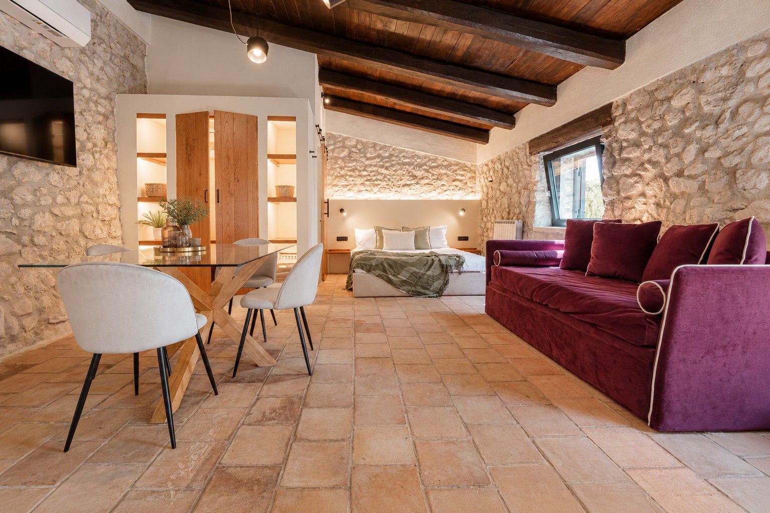 A cozy living area with a stone wall, a wooden ceiling, a purple sofa, and a dining table with chairs.