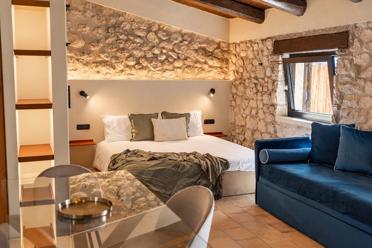 A bedroom with a stone wall, a bed with white bedding, a blue sofa, and a glass table with a metal base.
