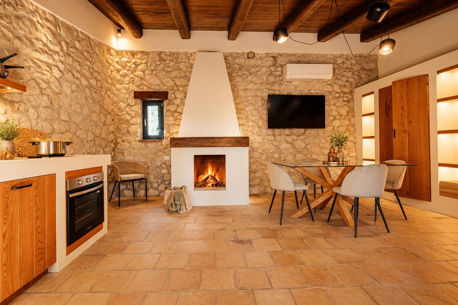 A stone-walled kitchen with a fireplace, wooden ceiling beams, and a dining area with chairs and a table.