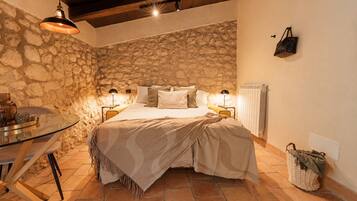 A bedroom with a stone wall, a bed with a beige blanket, two bedside lamps, a small table with a lamp, and a wicker basket.