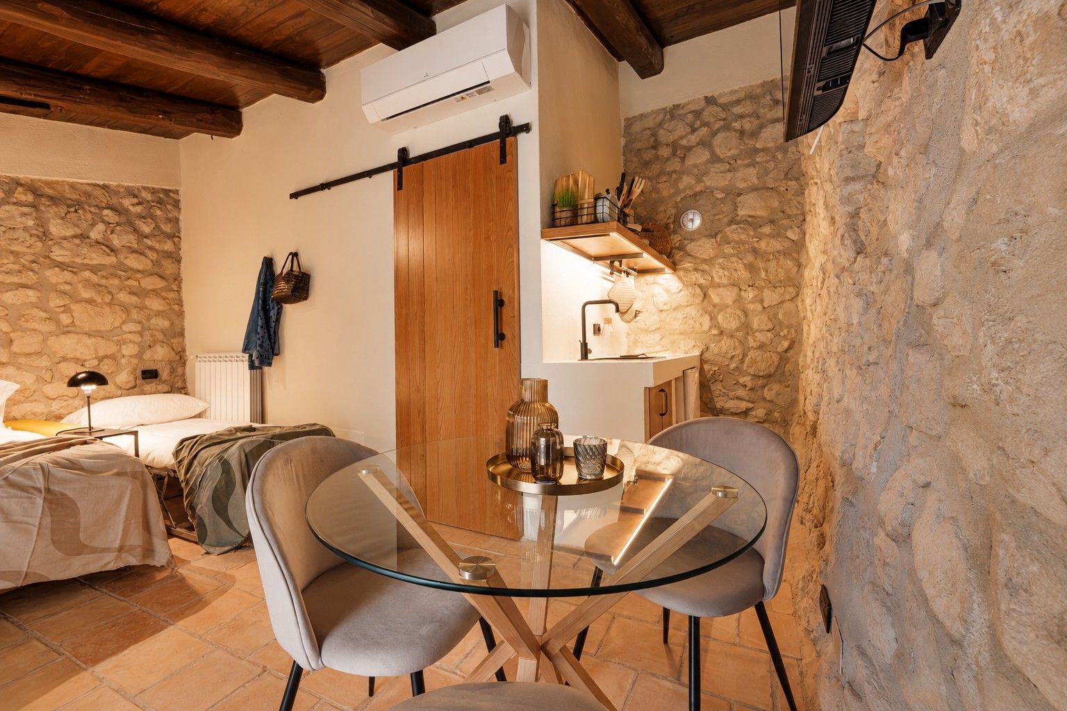 Una stanza accogliente con parete in pietra, porta in legno, un letto, un tavolo con sedie e una pianta pensile.