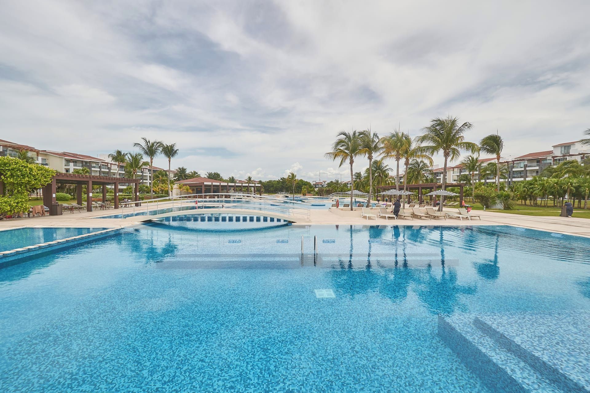 A large outdoor swimming pool with multiple lanes, surrounded by palm trees and lounge chairs, with buildings in the background.