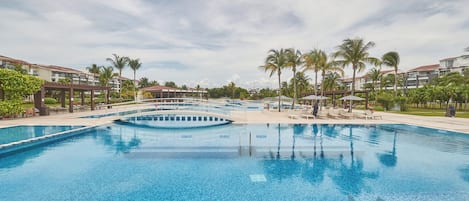 A large outdoor swimming pool with multiple lanes, surrounded by palm trees and lounge chairs, with buildings in the background.