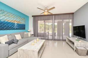 A modern living room with a grey sofa, a wooden coffee table, a TV on a wooden stand, and a ceiling fan. - Mareazul luxury condo | Beach & Resort Pools (Playa del Carmen)