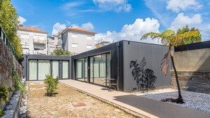 A modern building with large glass windows, a metal exterior, and a small garden area with a tree and gravel path. - GuestReady - PoetikBlue - Studio 4 (Porto)